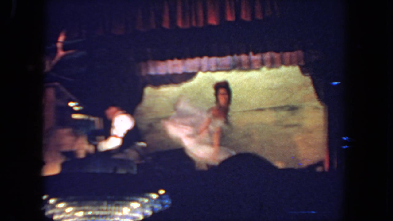 Woman in a light dress performs on a dim stage beside a seated keyboardist.