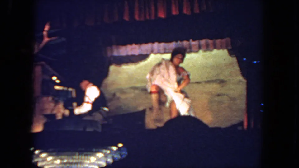 The performer turns near the center of a dimly lit San Diego stage, with a keyboardist seated to the side.