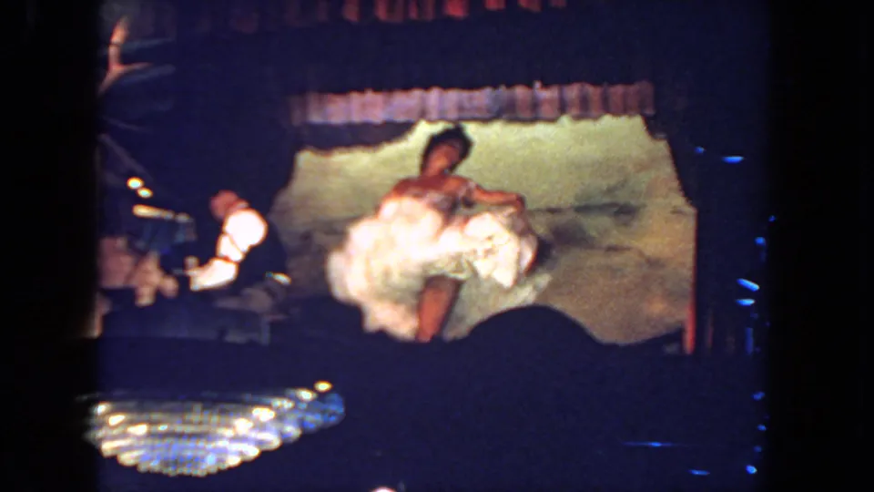 1962 San Diego home-movie frame showing a bride dancing energetically on stage while a tuxedoed pianist plays nearby.