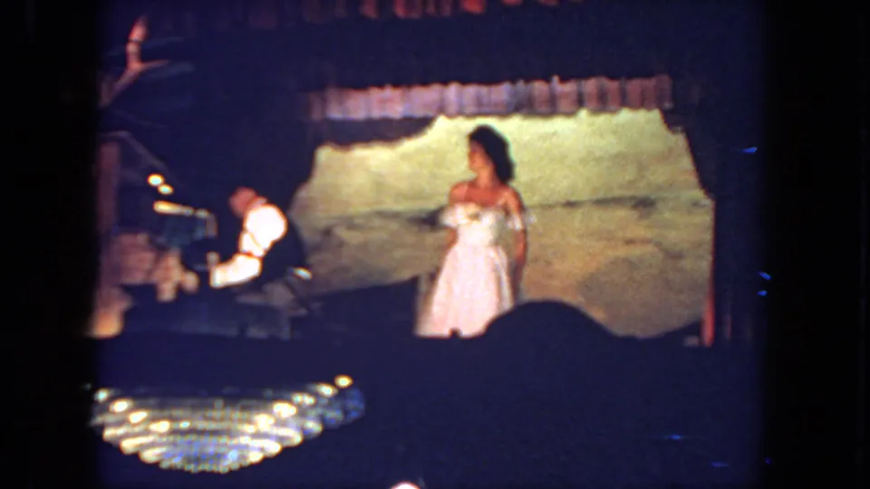 1962 San Diego home-movie frame of a bride near a pianist, filmed from the audience with chandelier light in front.