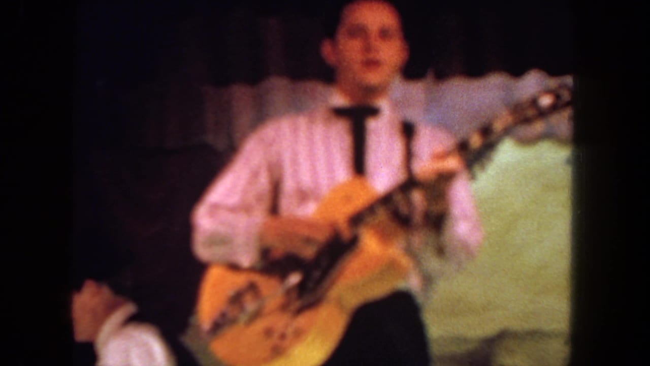 In supported 1962 San Diego footage, a man in a striped shirt and tie plays a yellow electric guitar under dim stage lighting.