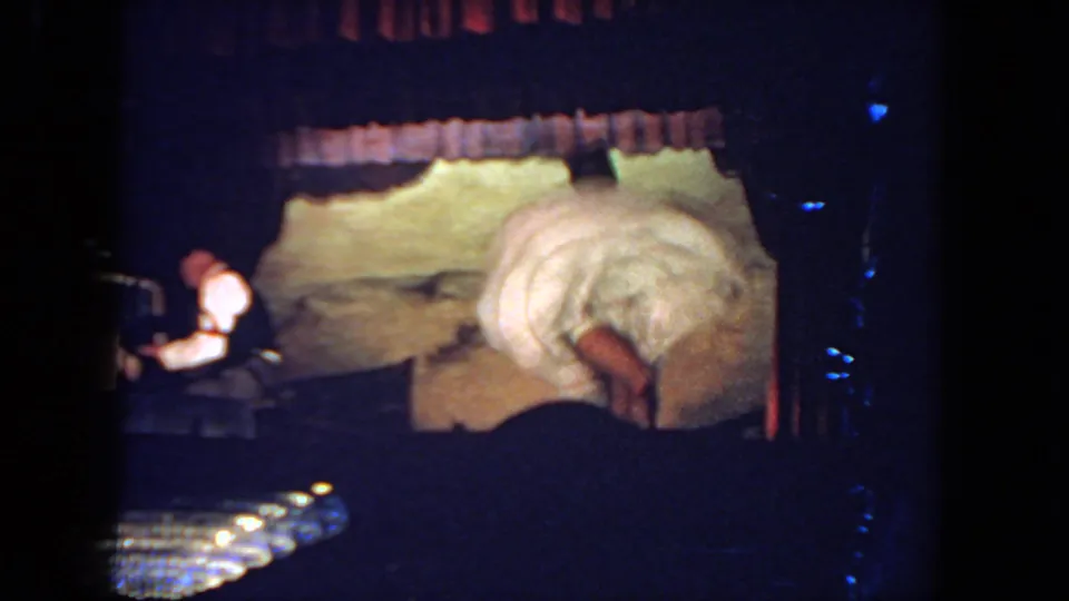 A woman in a white dance costume bends forward on stage while a pianist in a white shirt plays at the side.