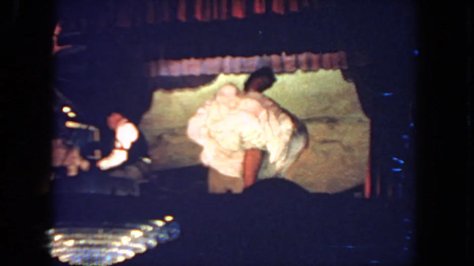 The dancer turns in a white costume on a dimly lit stage, with the pianist and backdrop visible in the same 1962 San Diego performance.