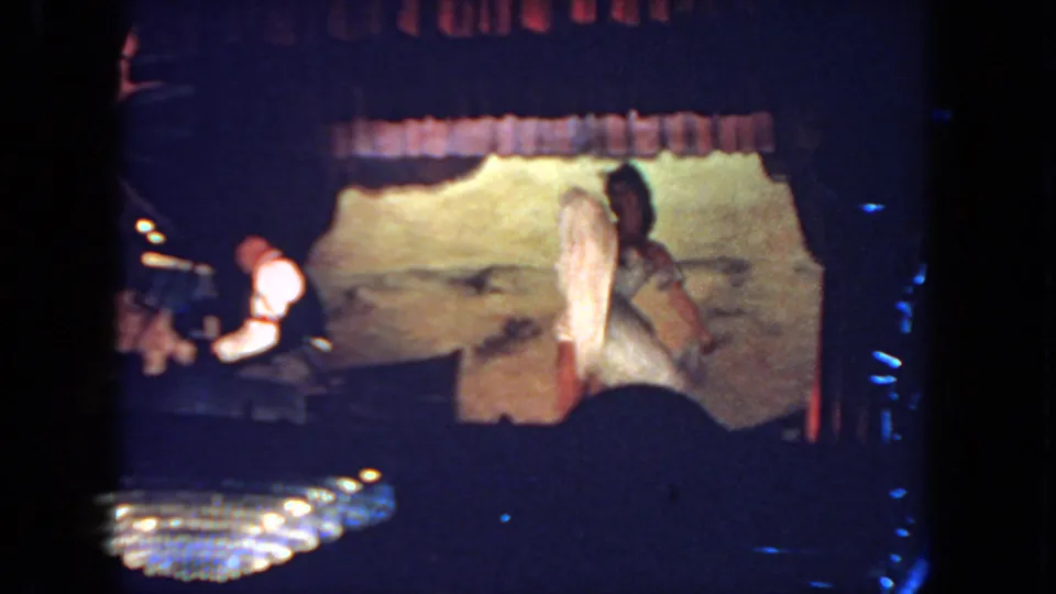 A dancer in a white costume lifts her leg during the routine as the pianist plays at stage left under low theatrical lighting.