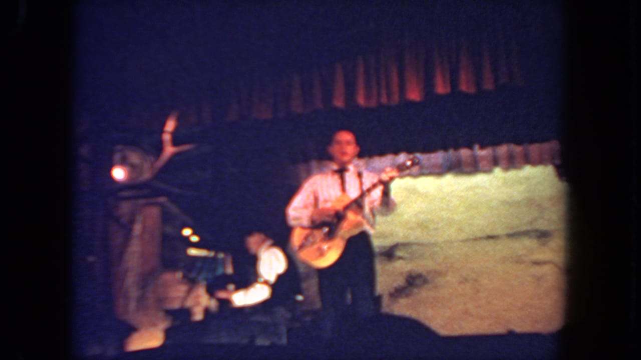 Wide stage view of a man playing electric guitar with another musician seated nearby.