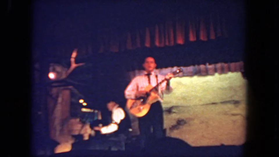 1962 San Diego home-movie frame showing a man on stage with an electric guitar, microphone, stage light, curtain, and seated bandmate.