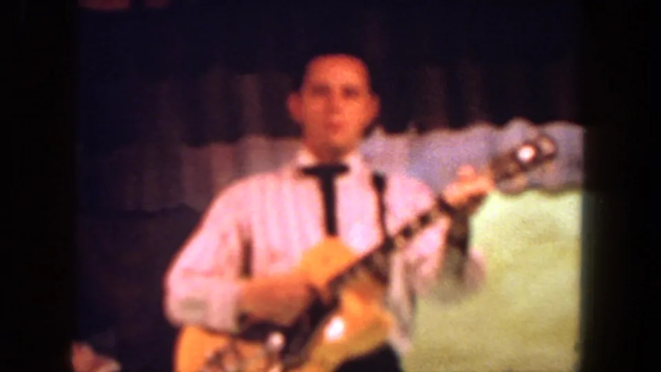 Close 8mm frame of the guitarist in a striped shirt and tie, standing before a dark curtain and pale stage backdrop.