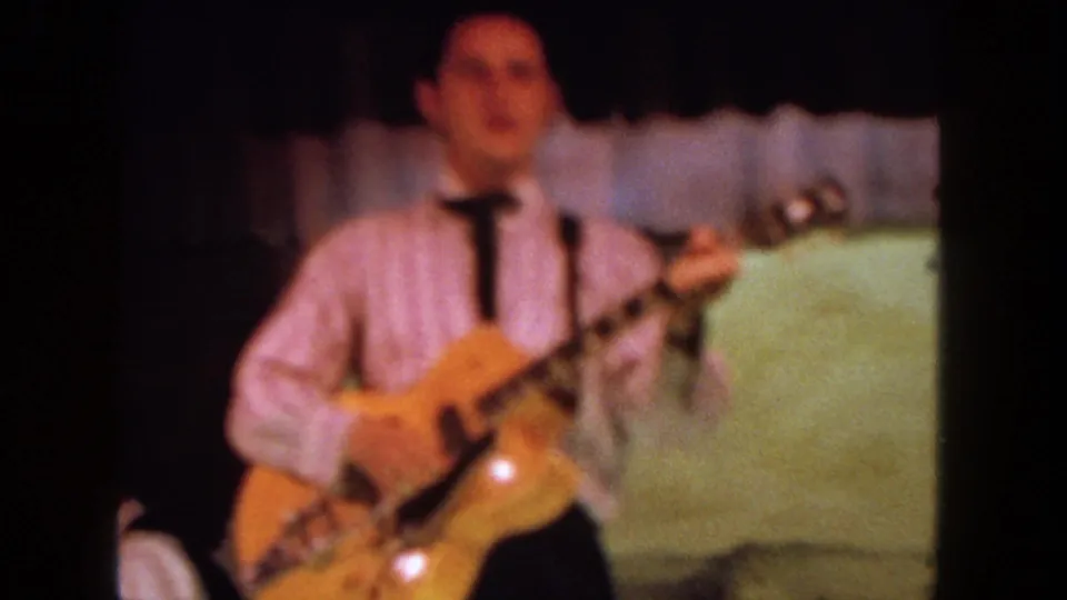 Soft-focus 8mm view of the guitarist and sunburst electric guitar, with the stage backdrop and part of another performer at the edge.