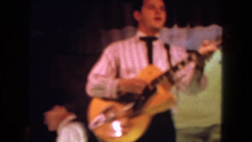 1962 San Diego frame showing the guitarist mid-performance, microphone nearby, with a seated bandmate partly visible against the stage backdrop.