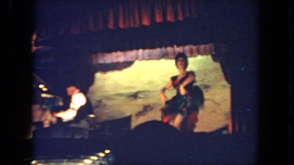In 1962 San Diego footage, a woman dances on a dim stage while a man sits at a piano to the left.