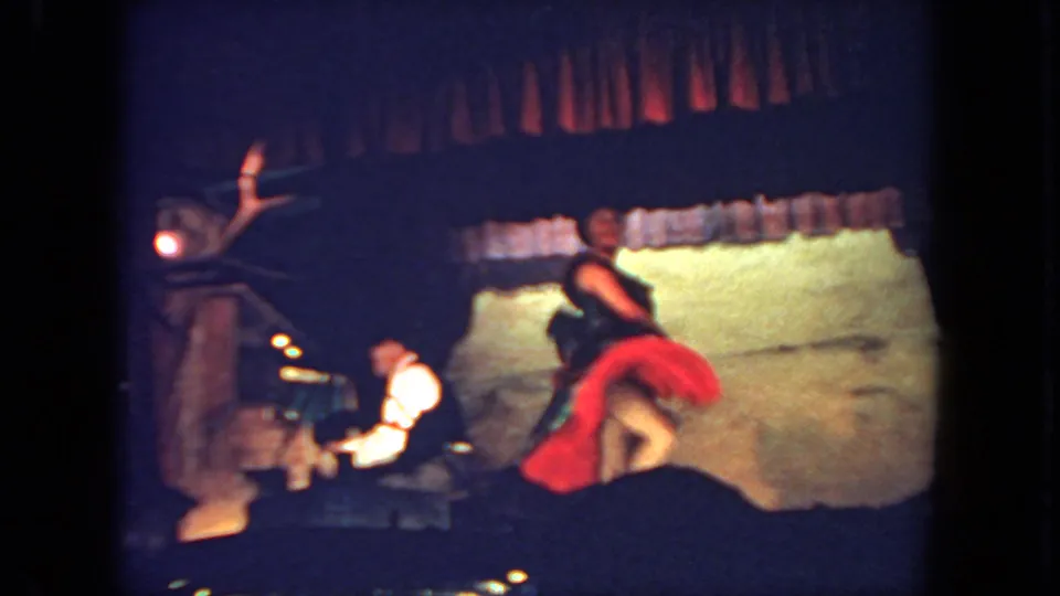 The dancer moves low or turns mid-step on the stage, with the pianist seated at the piano on the left.