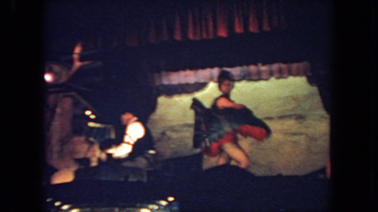 Woman in red and dark dress dances on stage as a man plays piano beside her.