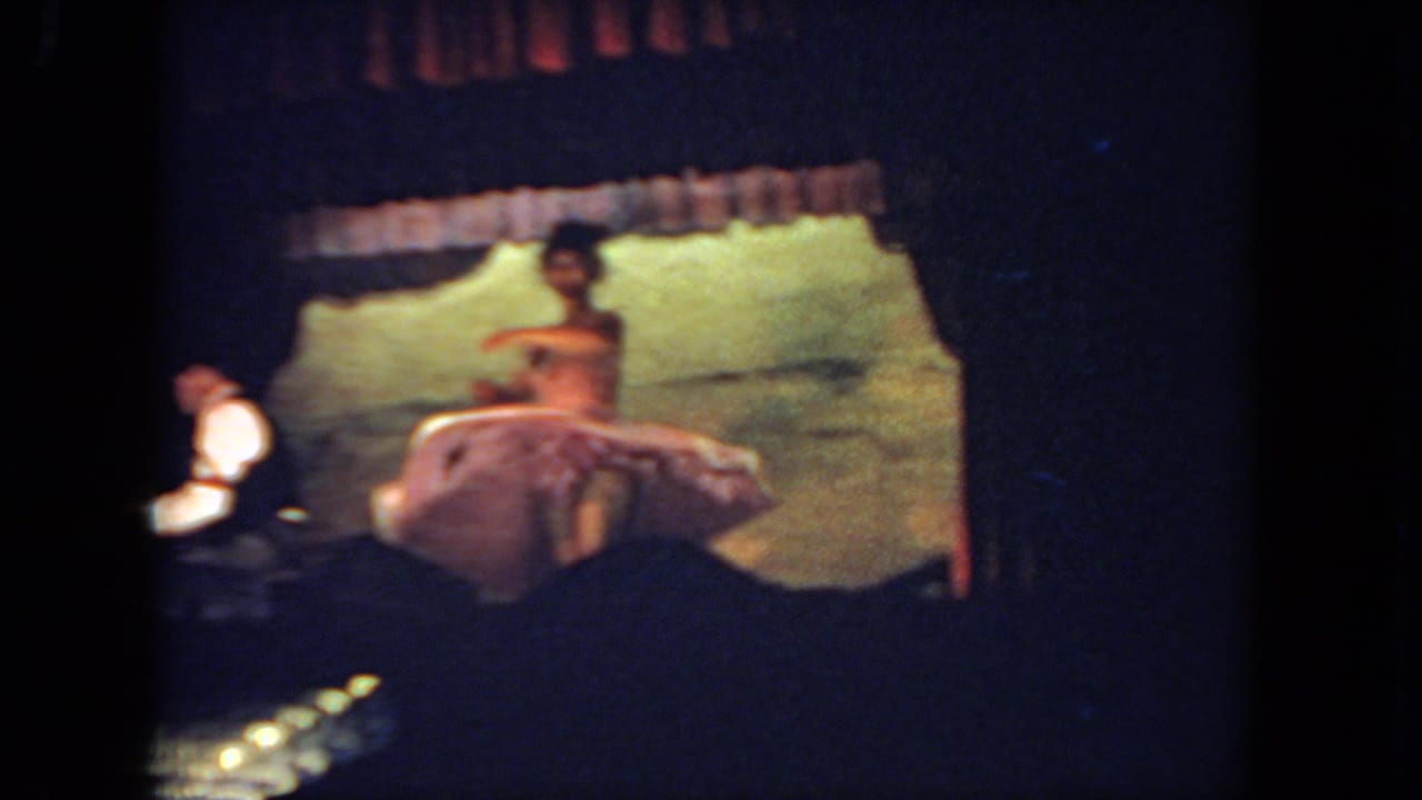 Dancer in a pale dress performs on stage while a pianist sits at the left and audience silhouettes fill the foreground.
