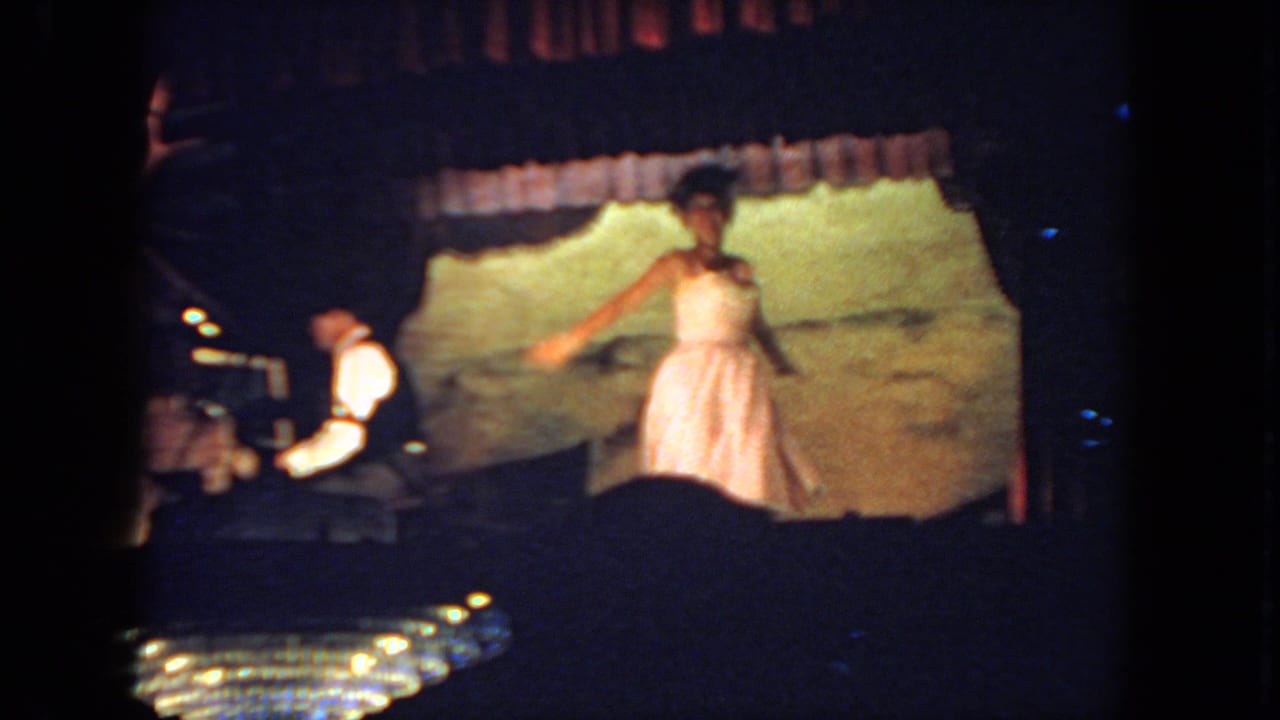 Woman in a pale dress dancing on a dim stage beside a seated pianist.