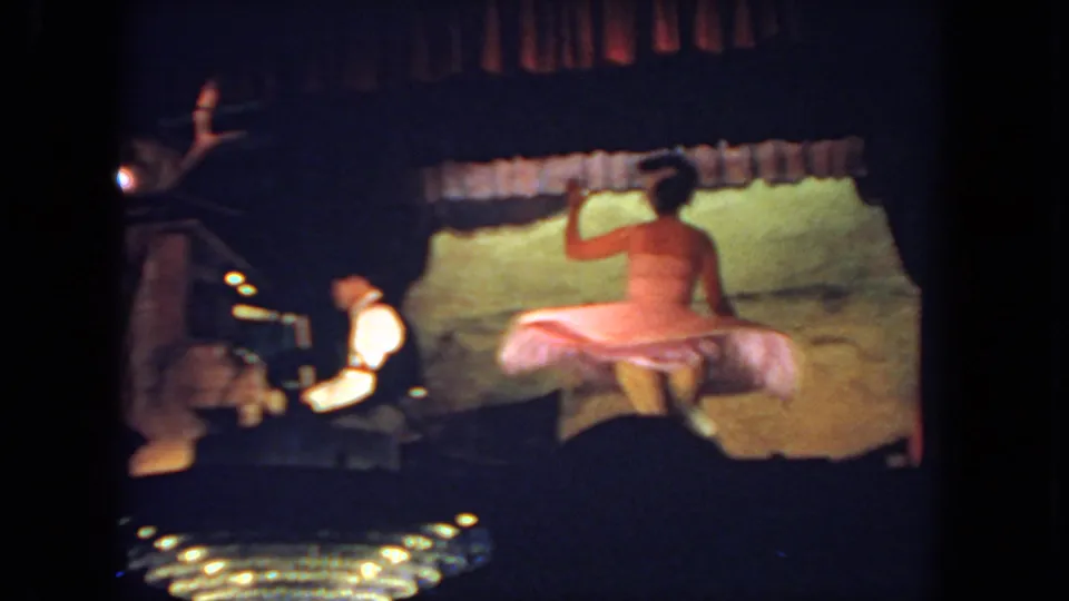 A woman spins in a pale pink dress on a dim 1962 San Diego stage, with the seated pianist visible at the left.