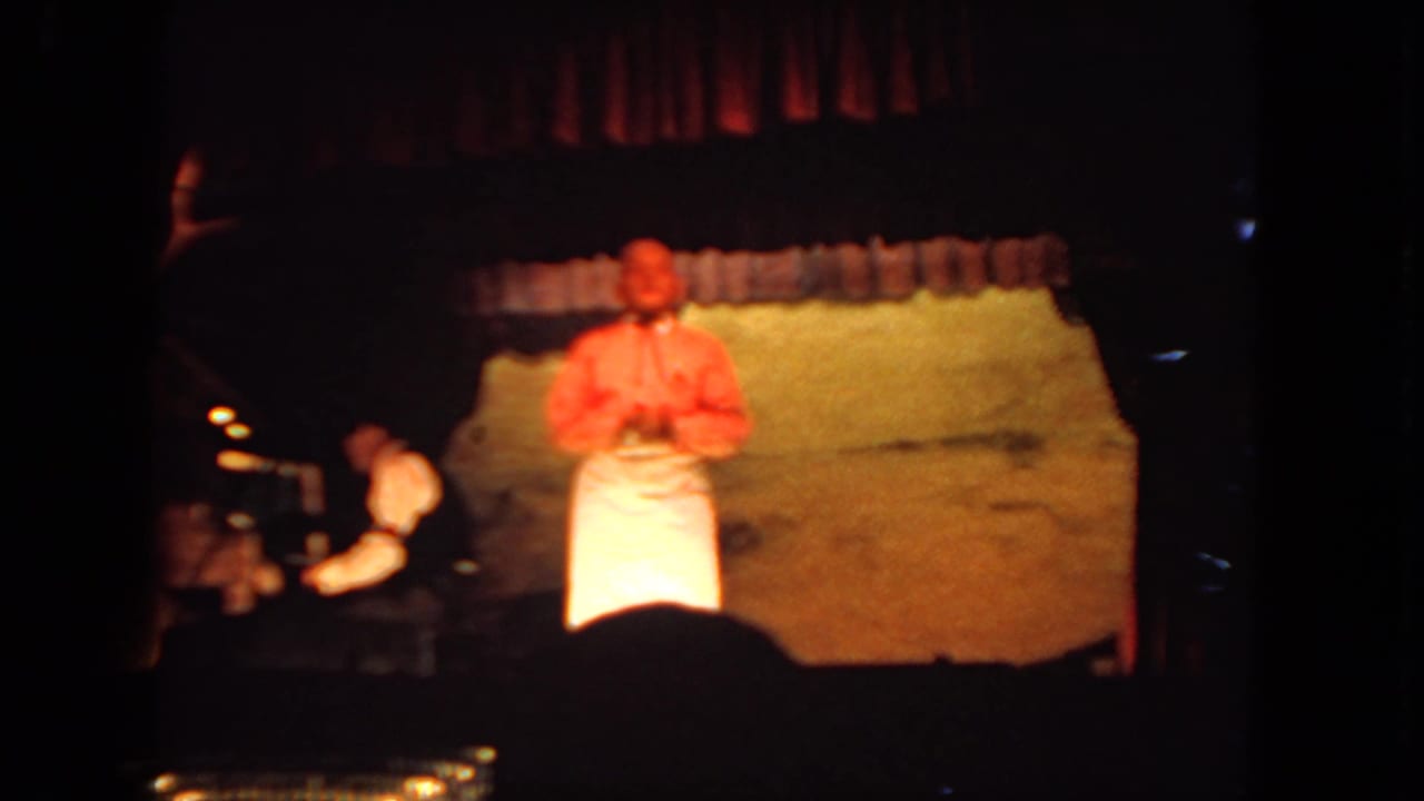 Man in red shirt and white apron stands on a dim stage beside a seated pianist.