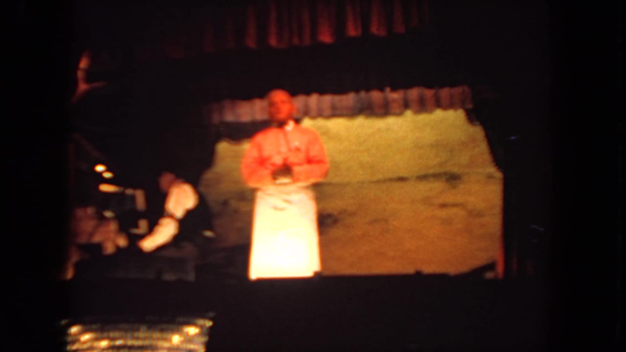 Presenter stands under warm stage light with a pianist partly visible at left.