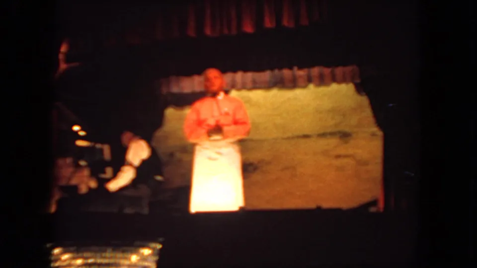 1962 San Diego stage-performance frame with a presenter in red and white under warm light and piano accompaniment at left.