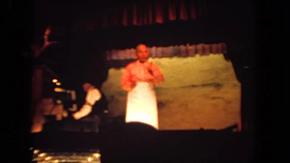 Home-movie frame from a 1962 San Diego stage performance, with a man presenting near center and a pianist in shadow at left.