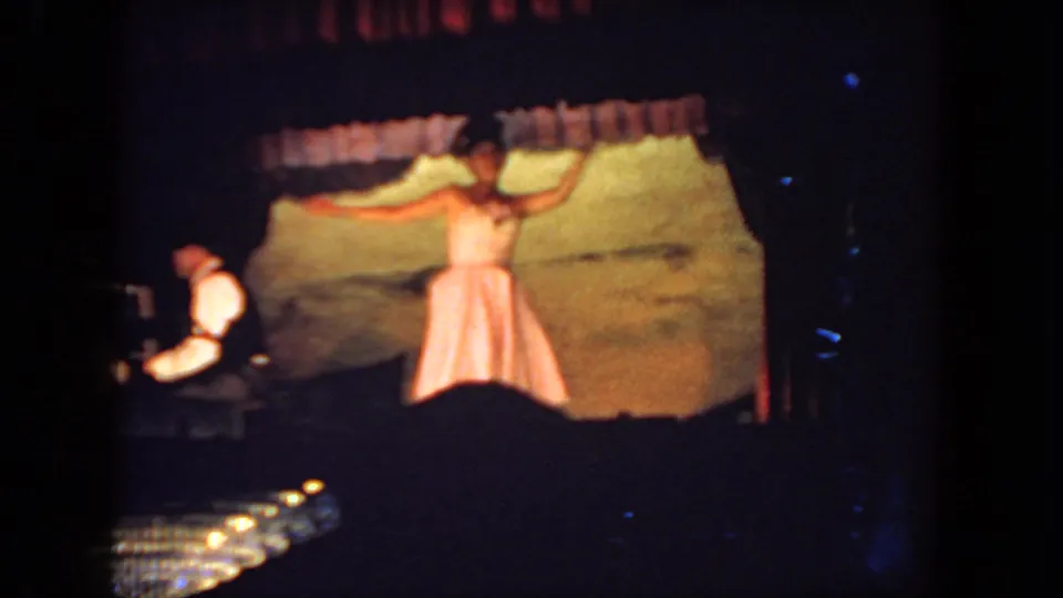 1962 San Diego home-movie frame showing a woman dancing on a curtained stage with a pianist accompanying at left.