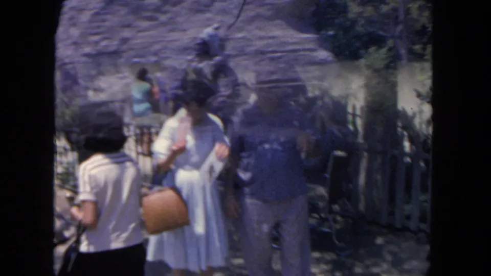 1962 San Diego home-movie frame of visitors on a sunny fenced path, including a woman in a white dress and other adults nearby.