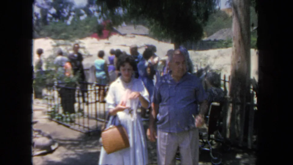 1962 San Diego visitor-shot frame showing a couple walking through a crowded outdoor attraction with trees and fencing behind them.