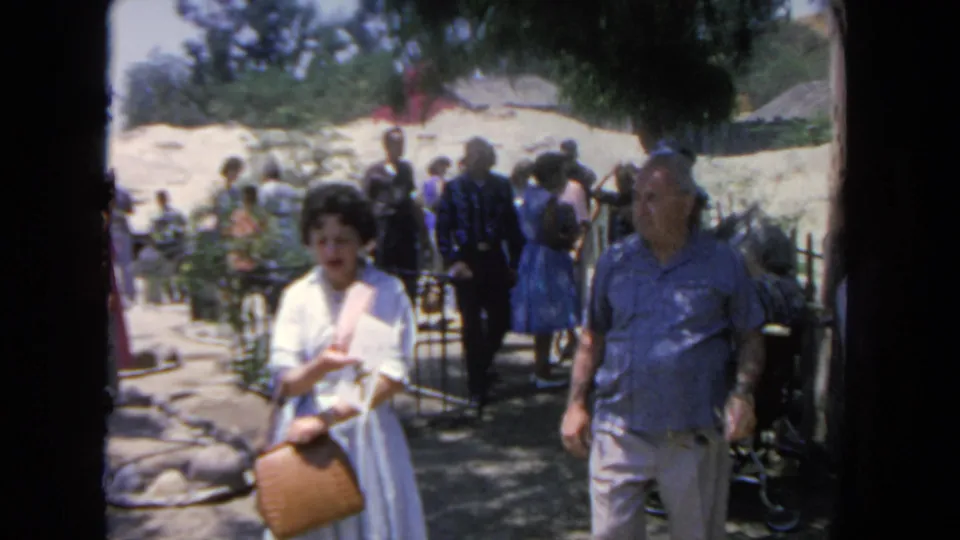 1962 San Diego home-movie frame of a woman with a tan purse and a man in a blue shirt walking through a busy outdoor walkway.