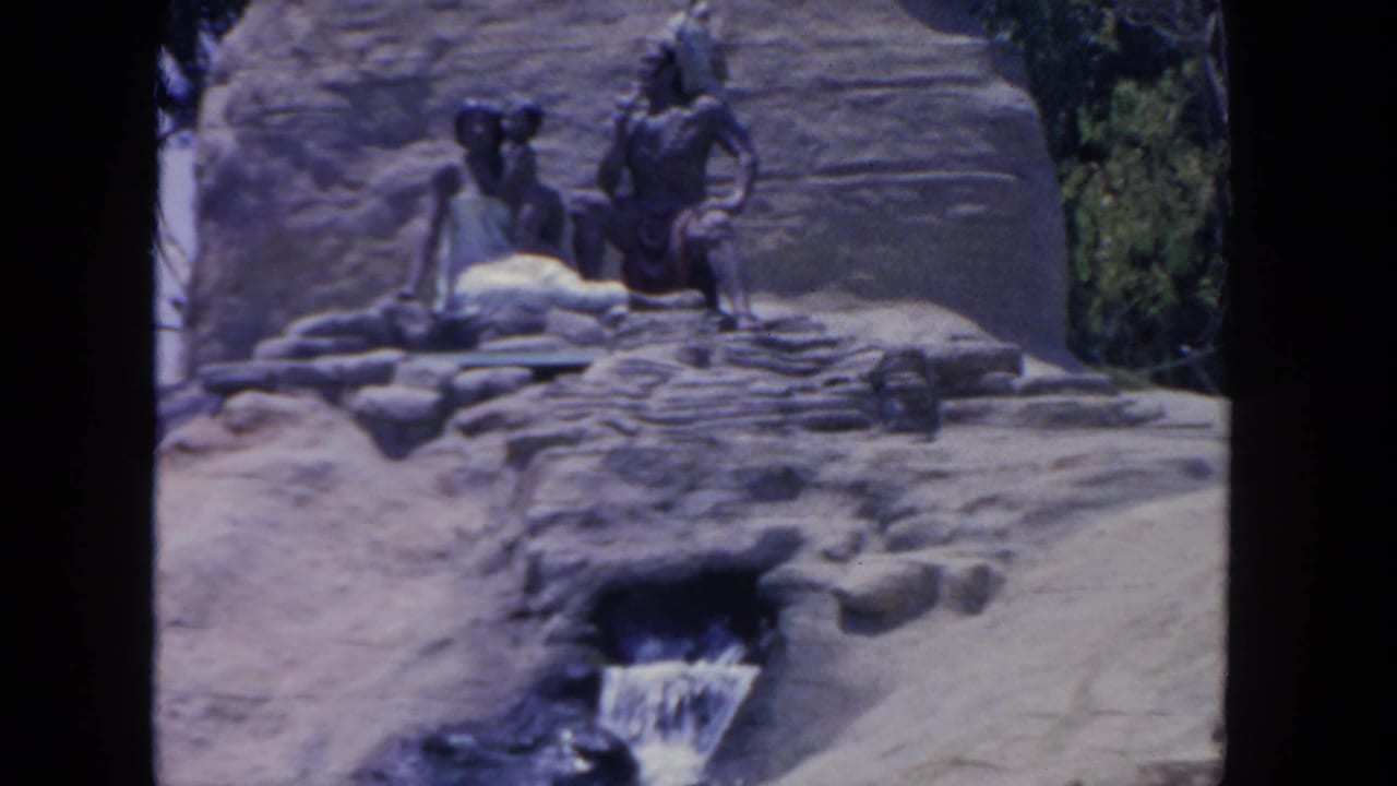 Visitor-shot 1962 San Diego zoo exhibit frame showing statues, terraced rocks, greenery, and a small artificial waterfall.