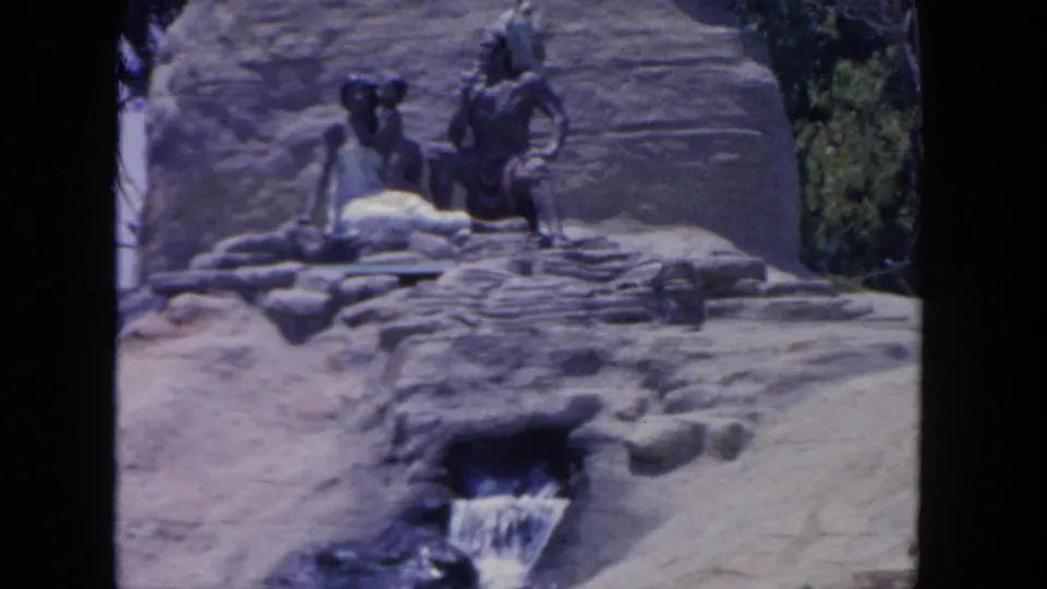 Visitor-shot 1962 San Diego zoo exhibit frame showing statues, terraced rocks, greenery, and a small artificial waterfall.