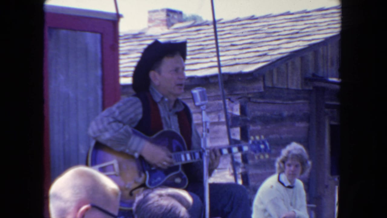 Man in cowboy hat plays electric guitar and sings into a microphone on an outdoor rustic stage.