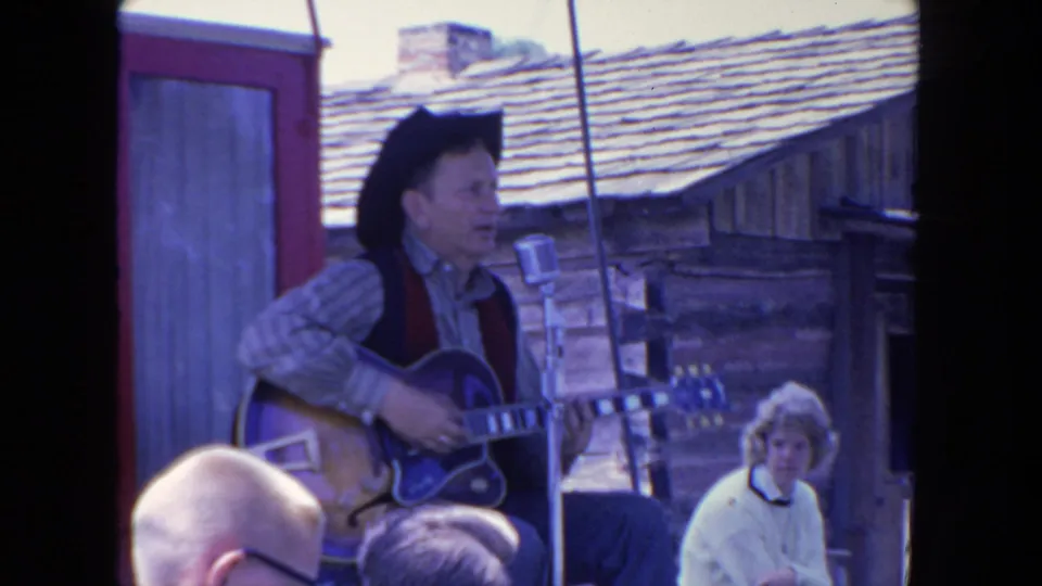 In San Diego, 1962, a man in western-style clothing performs with electric guitar and vintage microphone before seated spectators.
