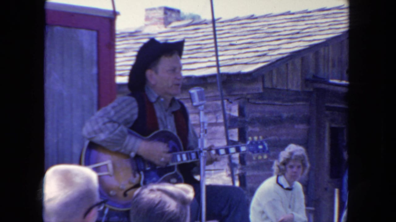Singer with electric guitar faces a microphone while audience heads fill the foreground.