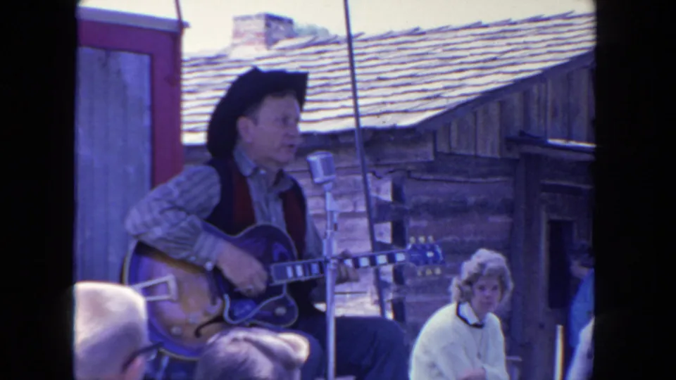 A western-dressed guitarist continues performing on the outdoor stage, framed by a cabin-like backdrop and nearby spectators.