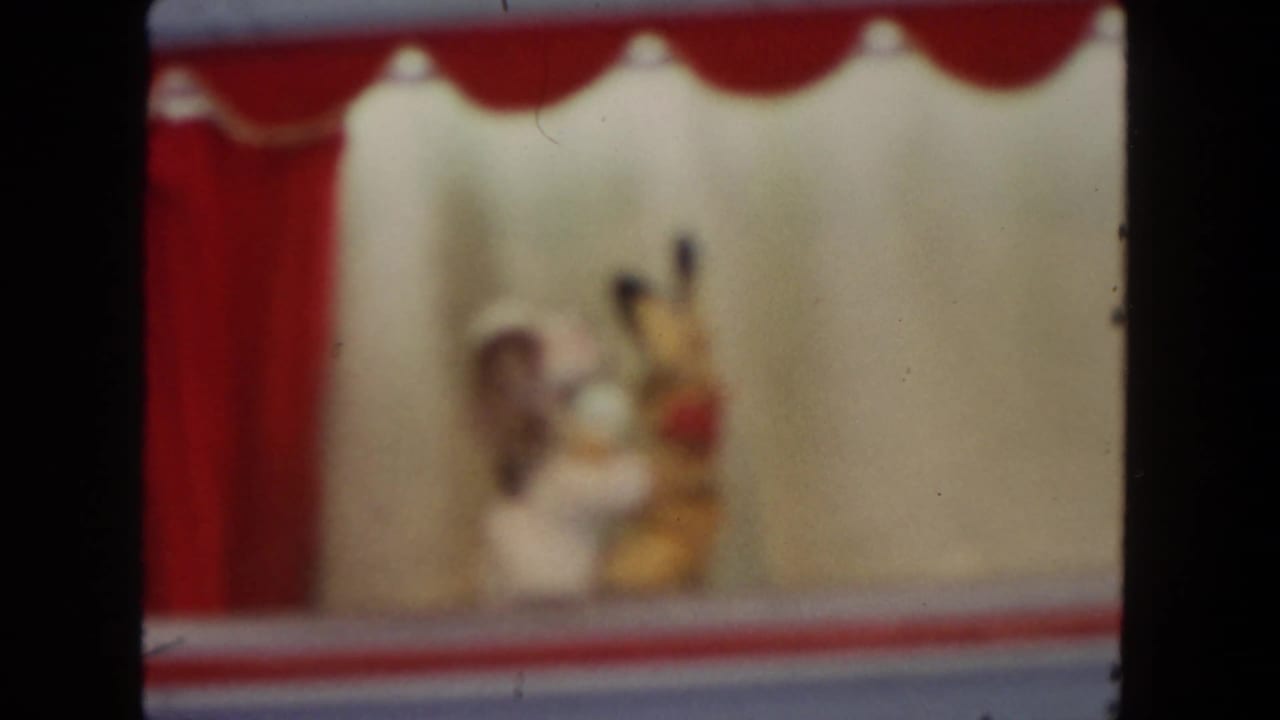 Soft-focus puppet stage with red curtain and two small figures behind a rail.