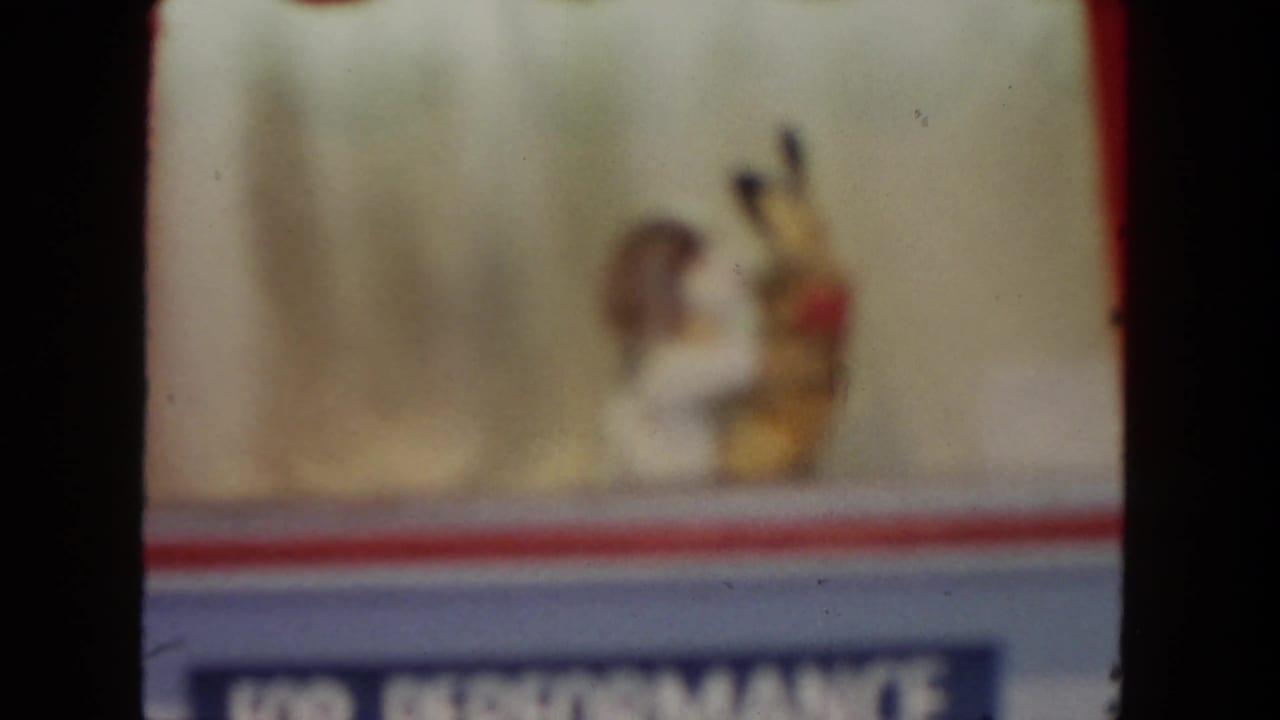 1962 San Diego 8mm frame with two soft-focus puppets performing above a blue lower sign area on a small stage.