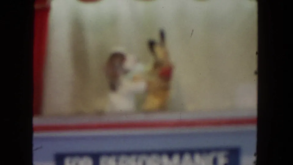 1962 San Diego 8mm frame showing the puppet figures in motion near the edge of a red curtain and stage front.
