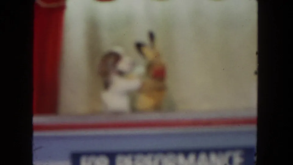 1962 San Diego 8mm frame of a white dog-like puppet and yellow rabbit-like puppet interacting on a small red-trimmed stage.