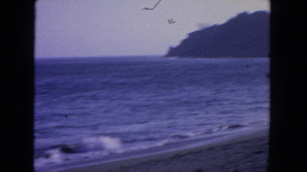 1962 San Diego 8mm frame showing surf on a sandy shoreline with open water and a dark headland across the horizon.