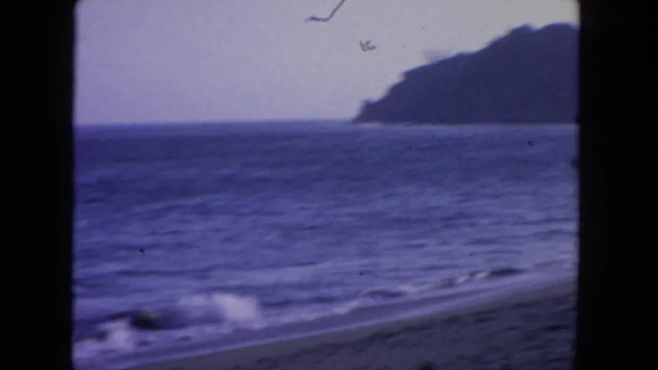 1962 San Diego 8mm frame showing surf on a sandy shoreline with open water and a dark headland across the horizon.