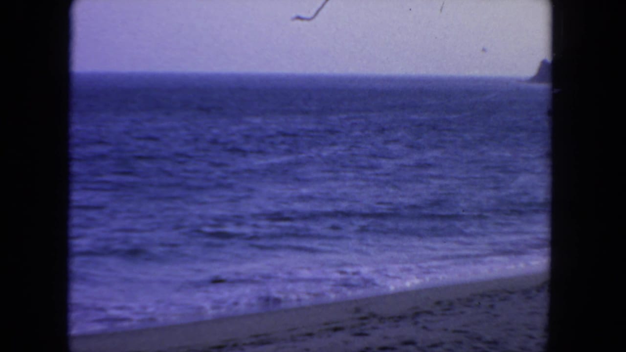 1962 San Diego coastal view on 8mm film, with rippled ocean, sloping beach, pale sky, and a small dark land edge at the far right.