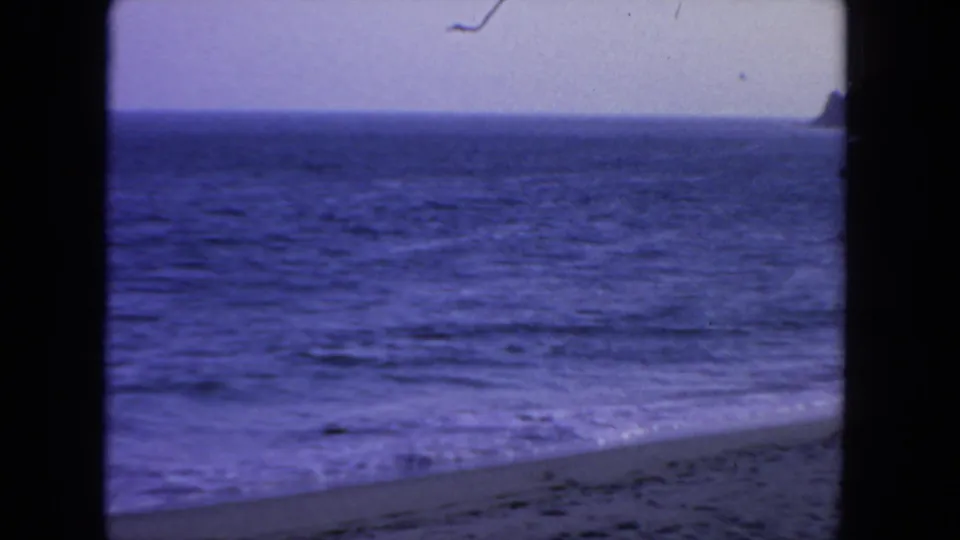 1962 San Diego coastal view on 8mm film, with rippled ocean, sloping beach, pale sky, and a small dark land edge at the far right.