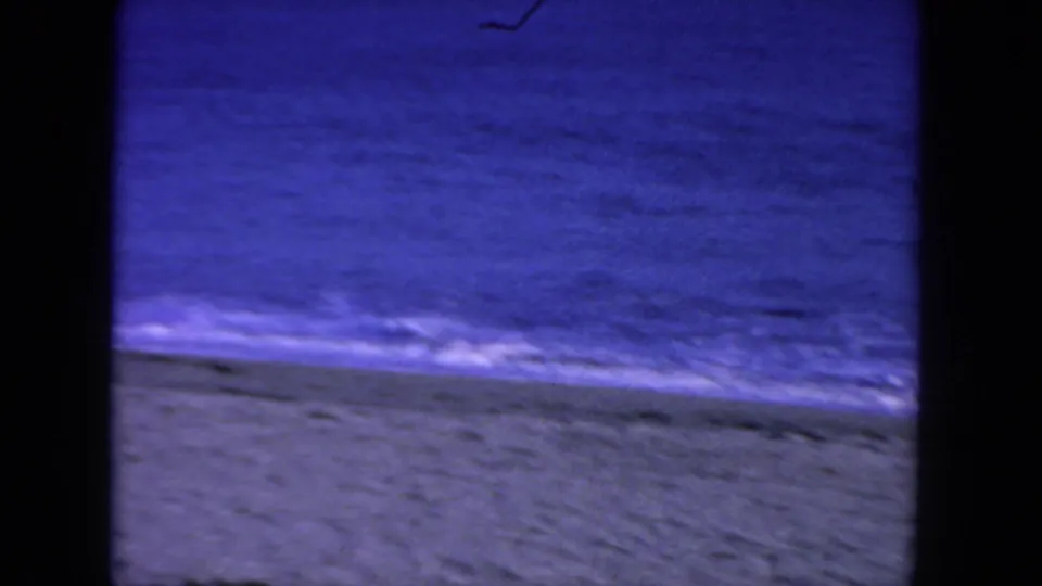 1962 San Diego beach frame with gentle surf, wet sand, and a broad expanse of blue-purple ocean on vintage 8mm film.