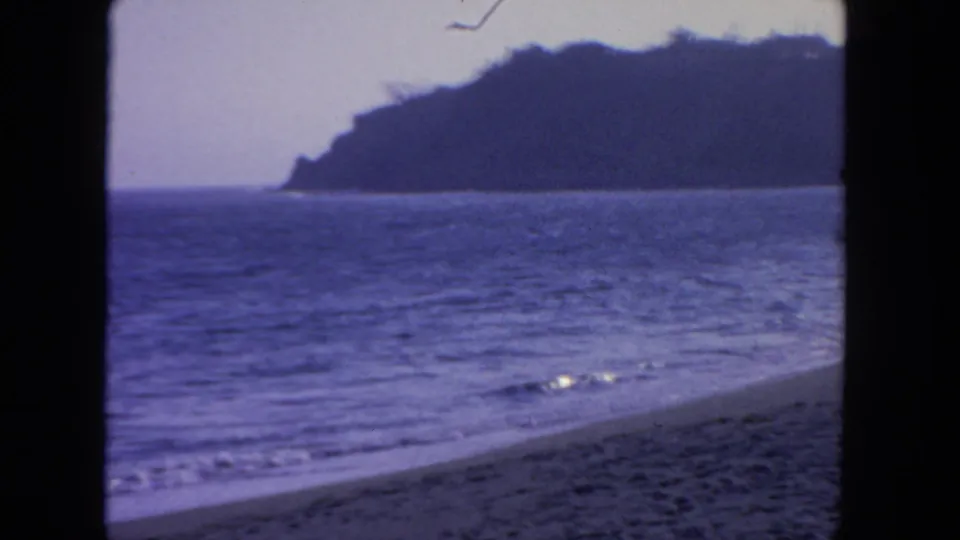 1962 San Diego 8mm frame showing calm surf, sandy foreground, and a distant bluff under soft light.
