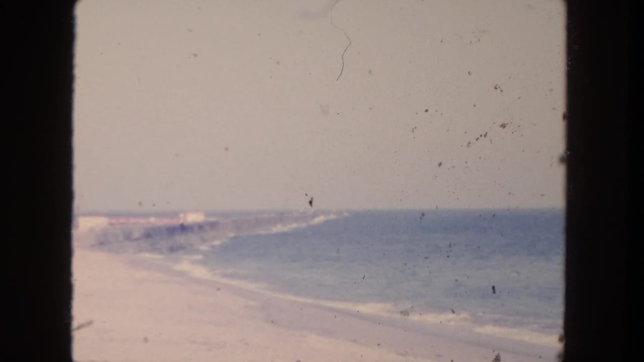 1962 San Diego beach view with gentle surf along pale sand under a hazy sky, seen in faded 8mm color.