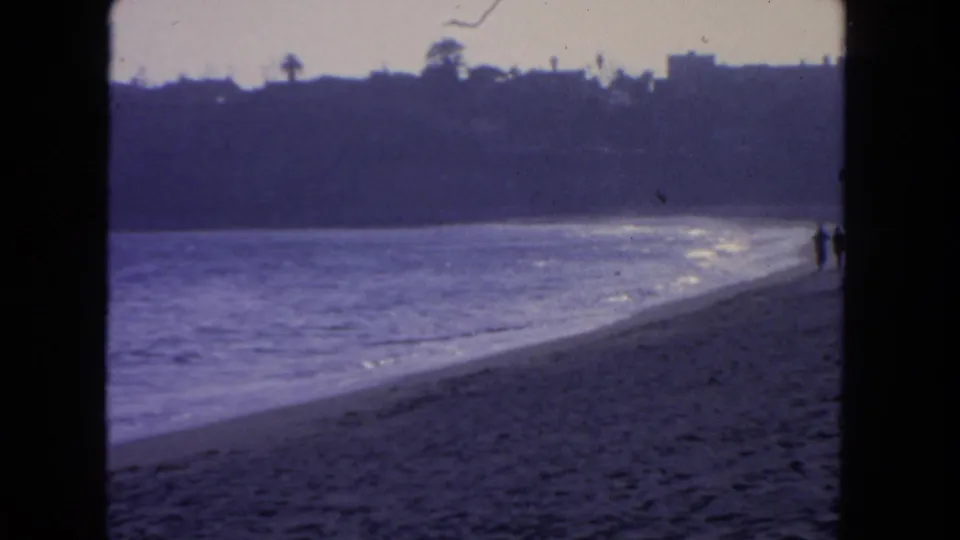 1962 San Diego 8mm beach frame showing surf, sand, distant hillside silhouettes, and two small walkers near the shoreline.
