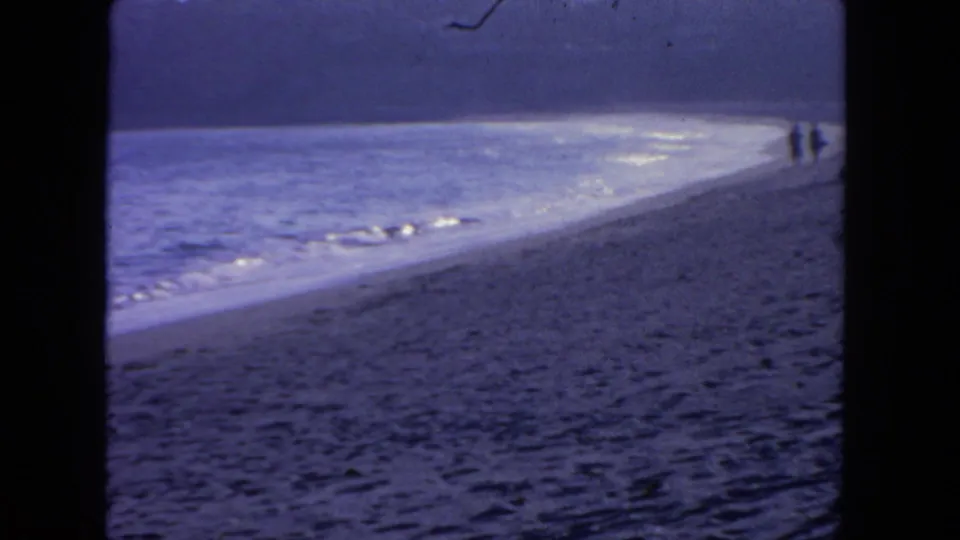 Faded 1962 San Diego beach view with foamy surf, textured sand, and two distant walkers along the waterline.