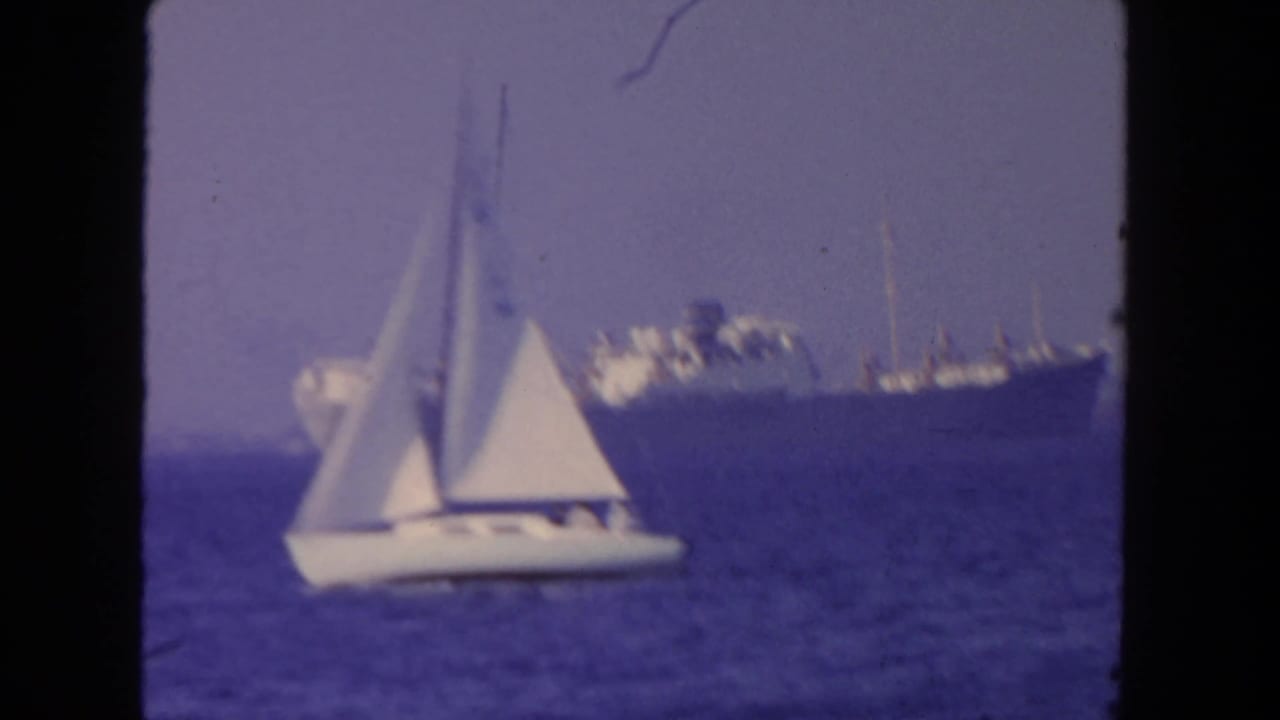 White sailboat on blue water with a large ship on the horizon.