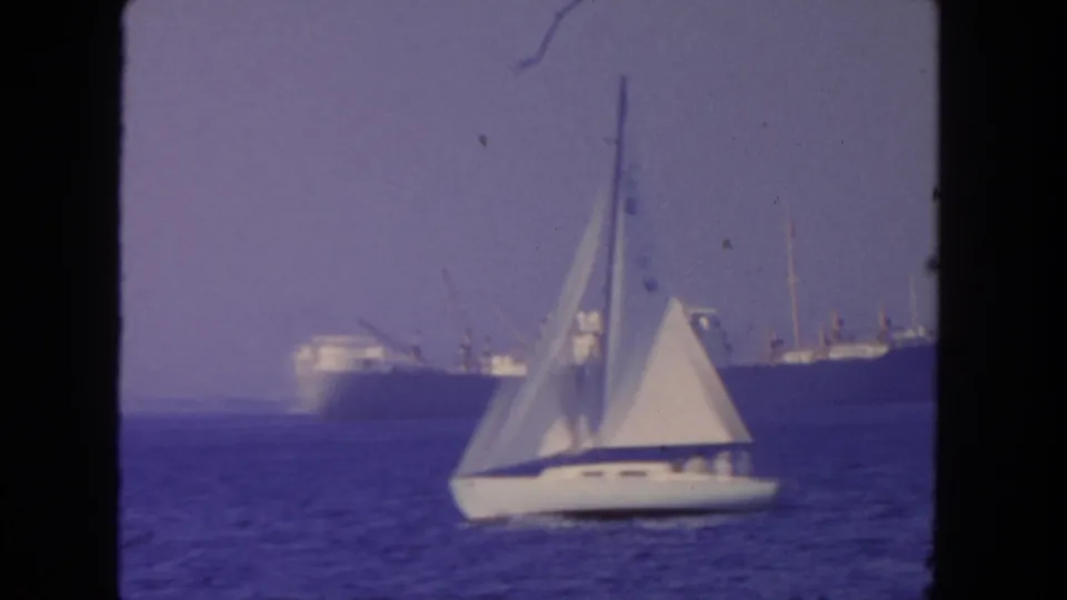1962 8mm frame of a white sailboat on the ocean with a large steamer blurred in the background.