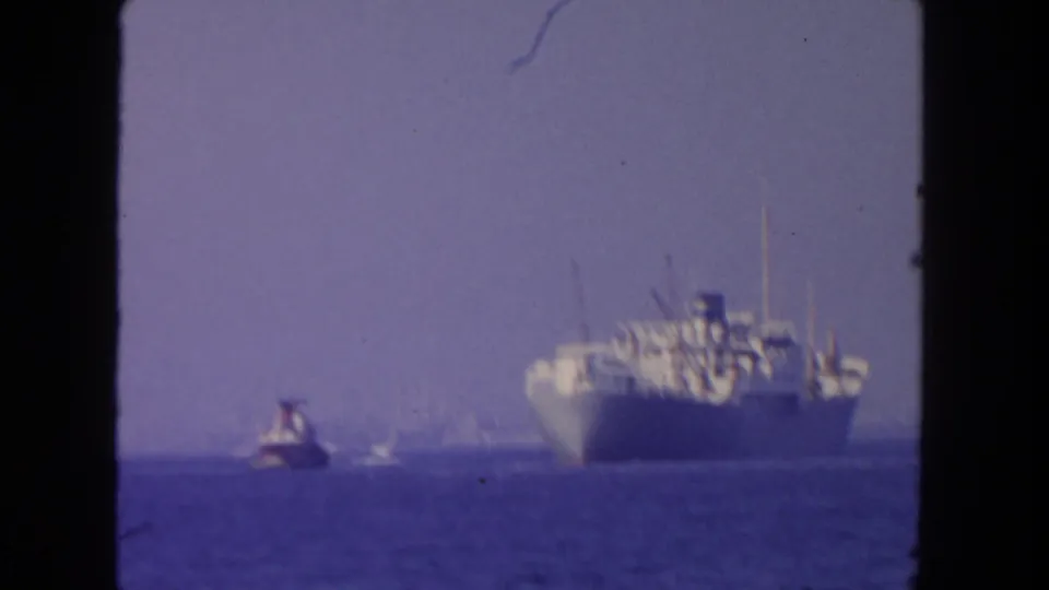1962 San Diego 8mm frame showing a large cargo ship at sea with a small tugboat to the left and a hazy horizon.