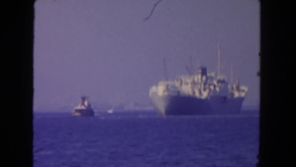 1962 San Diego 8mm frame with a cargo ship on the ocean, a tugboat ahead to the left, and a pale hazy sky.